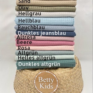 May include: A stack of waffle-weave fabric in various colors, including beige, olive green, and light blue. The fabric is labeled with German color names. A woven basket and a circular logo with the text "Betty Kids" are in the foreground.