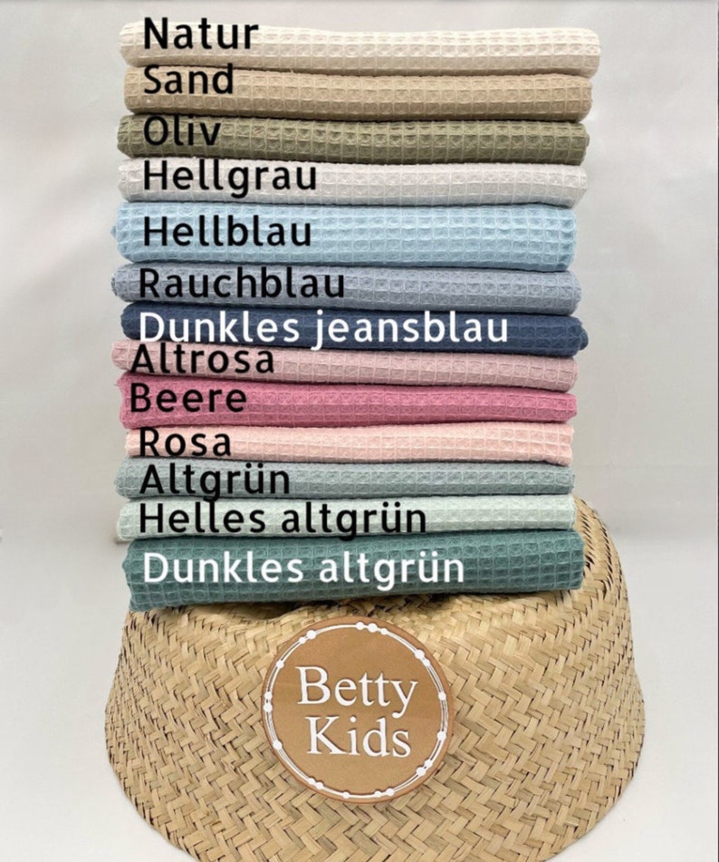 May include: A stack of waffle-weave fabric in various colors, including beige, olive green, and light blue. The fabric is labeled with German color names. A woven basket and a circular logo with the text "Betty Kids" are in the foreground.