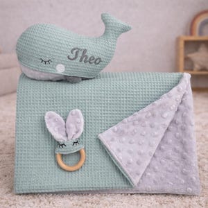 May include: A mint green baby blanket with a matching whale-shaped pillow embroidered with the name "Theo". A gray bunny-shaped teether with a wooden ring and a gray dotted blanket are also included. The items are on a light-colored carpet.