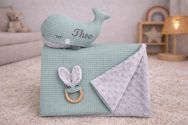 May include: A mint green baby blanket with a matching whale-shaped pillow embroidered with the name "Theo". A gray bunny-shaped teether with a wooden ring and a gray dotted blanket are also included. The items are on a light-colored carpet.