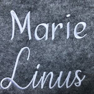 May include: A close-up of a gray felt material with the names "Marie" and "Linus" embroidered in white cursive script. The names are centered and take up most of the frame.