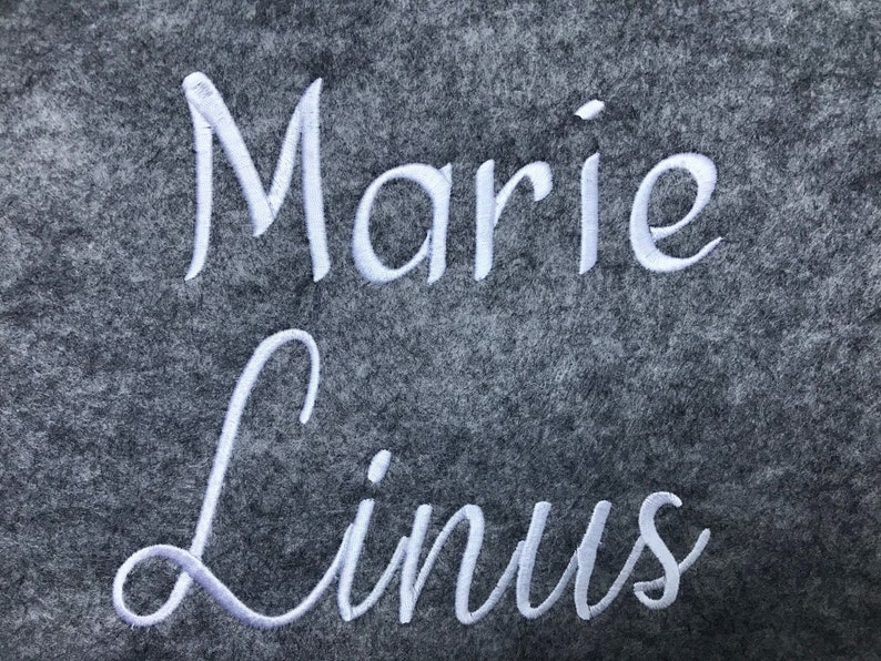 May include: A close-up of a gray felt material with the names "Marie" and "Linus" embroidered in white cursive script. The names are centered and take up most of the frame.