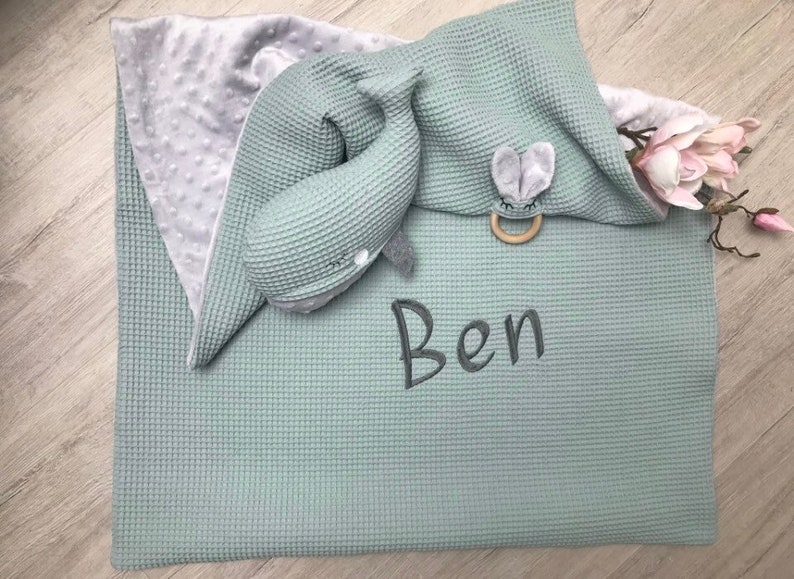 May include: A soft, mint green baby blanket with a waffle weave texture. A plush whale toy and a bunny-shaped teether rest on the blanket. The name "Ben" is embroidered on the blanket in gray thread. A gray, dimpled blanket is visible.
