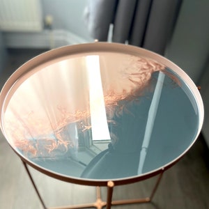 Puede incluir: Una mesa auxiliar redonda con un marco de metal de color cobre y una tapa circular de resina. La tapa es un diseño de dos tonos con un color rosa claro y azul grisáceo.