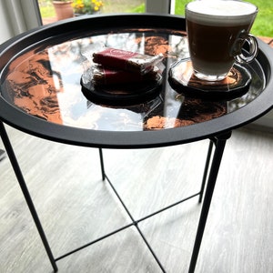 Puede incluir: Una mesa auxiliar negra con una tapa redonda de resina epoxi negra y cobre. La mesa tiene una base en forma de cruz y una taza de café y un paquete de galletas están sobre la mesa.