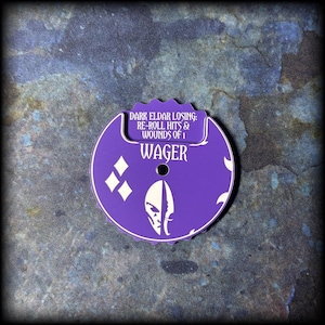 Könnte beinhalten: Ein lila Spiel-Token mit weißem Text und Grafiken. Der Text lautet "DARK ELDAR LOSING: RE-ROLL HITS & WOUNDS OF 1" und "WAGER". Der Token hat ein kreisförmiges Design mit einer gezackten Oberseite und einem Loch in der Mitte.
