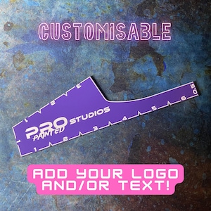 May include: A purple, customisable stencil with white text that reads "PRO STUDIOS PAINTED". The stencil has measurements from 0.5 to 6 inches (approximately 15 cm). The text "ADD YOUR LOGO AND/OR TEXT!" is in a pink box.