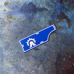 May include: A blue and white rectangular object featuring a sun and creature design. The object has measurements marked on it. The background is a mottled blue and grey. The measurements are in inches.