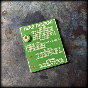May include: A green Hero Tracker card with white text and a circular emblem. The card details game instructions, including move, shoot, and charge actions. The emblem features a skull and a sun-like design. The card measures approximately 7.6 x 5 cm.