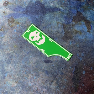 May include: A green sticker featuring a white skull design and the number "0.5". The sticker is rectangular with a curved bottom edge and a white border. The skull is a simplified, cartoon-like design.