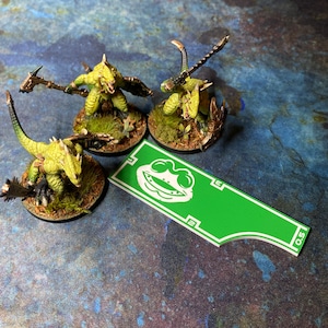 May include: Three miniature green lizard-like figures with weapons and detailed bases. A green and white game piece with a frog face graphic is also present. The figures are on a blue and green textured surface.