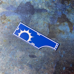 May include: A blue and white sticker featuring a sunburst design and the numbers "2", "3", and "0.5". The sticker has a distinctive shape with a curved edge. The background is a textured blue and green surface.
