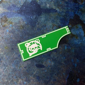 May include: A green and white die-cut sticker featuring a skull and floral design. The sticker has a rectangular shape with a curved edge and white border. The sticker has the numbers "2" and "0.5" printed on it.