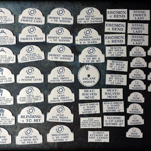 May include: A collection of white game tokens with dark blue text on a dark background. The tokens have various shapes, including arched tops and rectangles, with phrases like "Hurricane: Fights Last" and "Arcane Points".