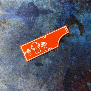 May include: An orange sticker featuring a white mushroom illustration. The sticker has a distinctive shape with rounded edges and a rectangular section with the text "E" and "0.5". The mushroom design includes three different mushroom shapes.