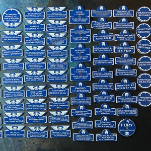 May include: A collection of blue and white game tokens with various text descriptions. The tokens are circular and rectangular, featuring phrases like "Oath," "Vengeance," and "Objective Secured." The tokens are for a tabletop game.