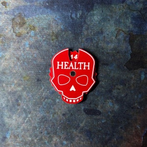 Op de afbeelding: Een rode, schedelvormige speltoken met het woord "HEALTH" in witte tekst. Het nummer "14" staat boven het woord "HEALTH". De token heeft twee ooggaten en een neusgat. De achtergrond is een gevlekt blauw en grijs.