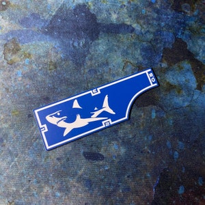 May include: A blue sticker with a white shark design. The sticker has the numbers 2, 3, and 0.5. The sticker is rectangular with a curved edge.