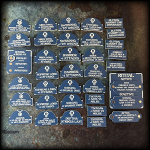 May include: A collection of navy blue game tokens with white text. The tokens have various phrases like "Supreme Lord +1 Attacks" and "Tsunami -1 Save vs Melee". Some tokens have a stylized emblem. The tokens are for a tabletop game.