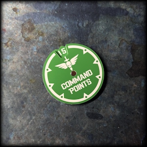 May include: A green and white circular game token with the text "COMMAND POINTS". The token has a notch at the top with the number "15". A winged skull and sword graphic is in the center.