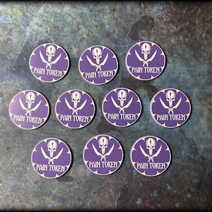 Könnte beinhalten: Elf runde, violette Spielmarken mit einem weißen Totenkopf mit gekreuzten Schwertern und dem Schriftzug "PAIN TOKEN". Die Spielmarken liegen auf einer dunklen, strukturierten Oberfläche. Jede Marke hat einen Durchmesser von etwa 3 cm.