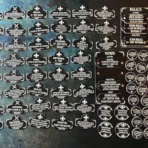 Pode incluir: Um conjunto de fichas de plástico preto e branco com vários textos e símbolos. As fichas são para um jogo de tabuleiro e incluem termos como "Zelote", "Mártir", "Votos", "Relíquias" e "Penitente".