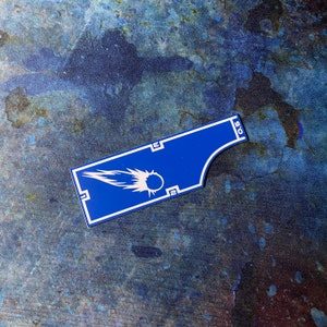 May include: A blue and white rectangular sticker featuring a comet graphic. The sticker has a curved edge and the number "0.5" printed on it. The sticker also has the numbers "2" and the letter "E" printed on it.