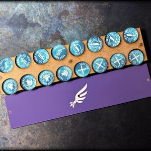 May include: A wooden game piece holder with 12 teal and white circular tokens. Each token has a different symbol and the words "TEST FOR" and another word. A purple card with a white logo is below the holder.