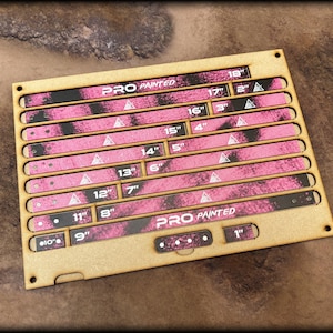 May include: A rectangular tan wooden board containing multiple removable measuring sticks with a pink and black camouflage pattern. The sticks are labeled with imperial measurements ranging from 1 inch to 18 inches. The text PRO PAINTED is printed on the board.