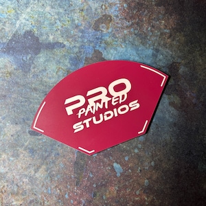 Könnte beinhalten: Ein fächerförmiges Schild in Burgunderrot mit dem Text "PRO PAINTED STUDIOS" in weißer, fetter, serifenloser Schrift. Das Schild hat weiße, eckige Randakzente. Der Hintergrund ist ein gesprenkelter Mix aus Blau-, Braun- und Grüntönen.