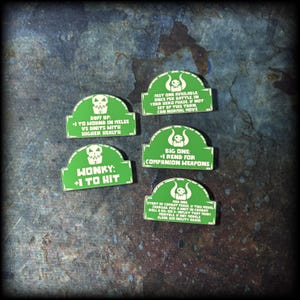 Peut inclure: Cinq jetons de jeu verts avec du texte blanc et des graphiques de crâne. Les jetons comportent diverses phrases, dont "DUFF UP", "WONKY" et "BIG ONE". Les jetons sont sur une surface sombre texturée.
