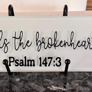 Op de afbeelding: Rechthoekige witte decoratieve plaat met zwarte tekst die luidt "he heals the brokenhearted" en "Psalm 147:3". De plaat heeft decoratieve zwarte hoekaccenten en wordt vastgehouden door een zwarte metalen standaard.