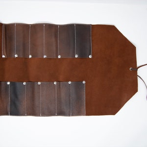 May include: A brown leather roll-up case with eight slots for holding tools or other items. The case has a tie closure and is made of a durable material.