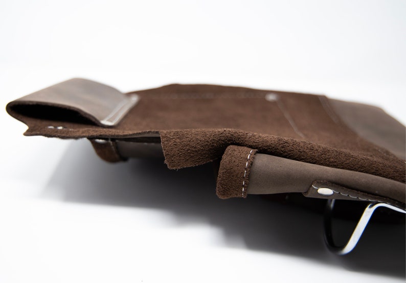 Puede incluir: Una bolsa de herramientas de cuero marr&oacute;n con una solapa y un asa de metal. La bolsa tiene un forro de gamuza y costuras blancas.
