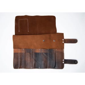 May include: A brown leather roll with six slots for holding tools. The roll has a flap that closes with a snap and a strap with a buckle.