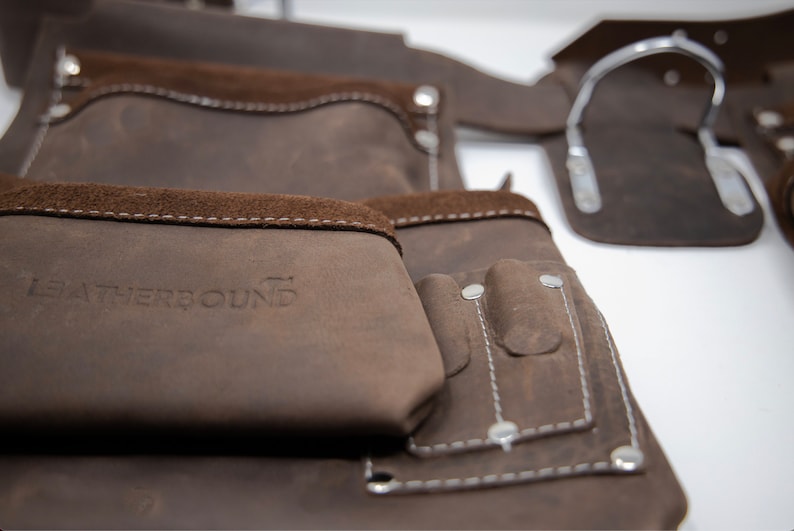 Puede incluir: Cintur&oacute;n de herramientas de cuero marr&oacute;n con m&uacute;ltiples bolsillos, incluido un bolsillo grande con el texto "LEATHERBOUND" en relieve.
