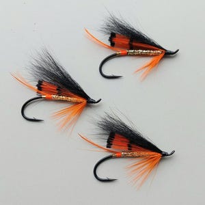 Puede incluir: Tres moscas de pesca con anzuelos negros, plumas negras y naranjas, e hilo dorado. Las moscas están dispuestas sobre una superficie gris claro, mostrando su intrincado diseño y colores vibrantes. Estas moscas están diseñadas para la pesca con mosca.