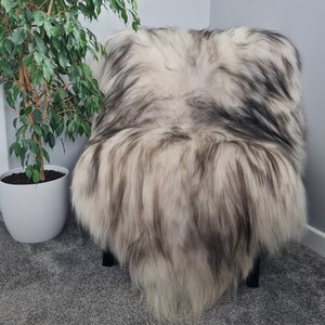 May include: A long-haired, faux fur throw draped over a chair, featuring a variegated colour pattern of cream, white, and dark charcoal grey streaks throughout the thick, shaggy texture of the material, creating a layered and voluminous appearance.