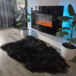 May include: A plush, black faux fur rug with a long, shaggy texture laid out on a light-colored wood floor. The rug has an irregular, organic shape and is positioned in front of a wall-mounted electric fireplace with a black glass panel.