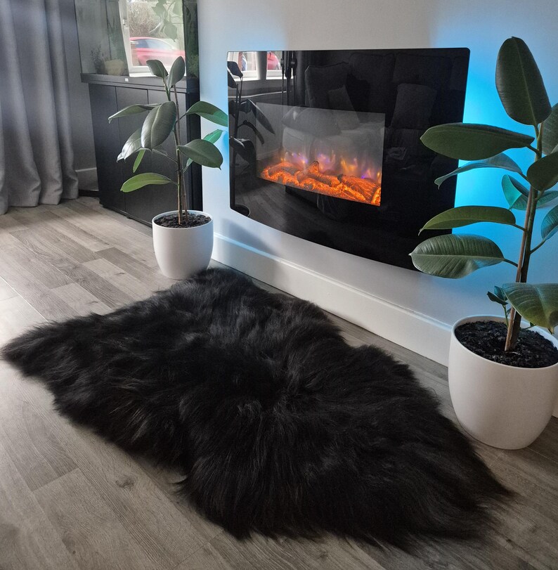 May include: A plush, black faux fur rug with a long, shaggy texture laid out on a light-colored wood floor. The rug has an irregular, organic shape and is positioned in front of a wall-mounted electric fireplace with a black glass panel.