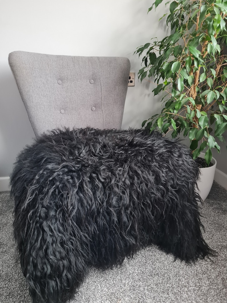 May include: A gray upholstered chair with four button tufts on the backrest, draped with a long-haired, dark charcoal black sheepskin rug that covers the seat and extends down the front of the chair frame.