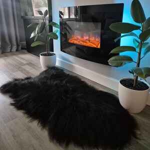 May include: A long-haired black sheepskin rug with a thick, shaggy texture. The rug is positioned on a light-colored wood-look floor, centered between two potted green plants and beneath a wall-mounted electric fireplace with a black glass front.