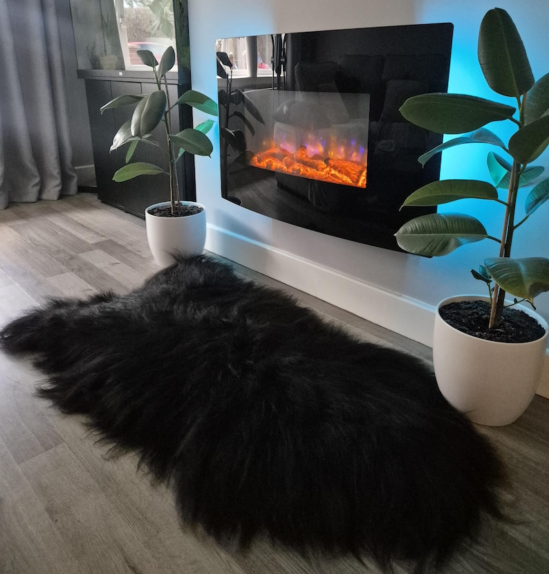 May include: A long-haired black sheepskin rug with a thick, shaggy texture. The rug is positioned on a light-colored wood-look floor, centered between two potted green plants and beneath a wall-mounted electric fireplace with a black glass front.