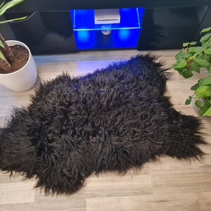 May include: A black sheepskin rug with long, curly, and dense fur. The rug has an irregular, natural shape with a thick, textured pile that covers the entire surface. The deep black color is consistent throughout the entire piece of animal hide.