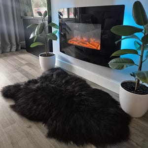 May include: A plush, black sheepskin rug with a thick, long-pile texture. The rug has an irregular, organic shape and is positioned on a light-colored wood floor, creating a soft contrast against the dark, dense fibers of the material.