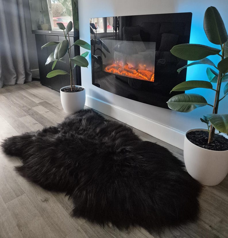 May include: A plush, black sheepskin rug with a thick, long-pile texture. The rug has an irregular, organic shape and is positioned on a light-colored wood floor, creating a soft contrast against the dark, dense fibers of the material.