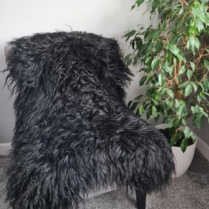 May include: A long-haired black sheepskin rug draped over a chair. The texture is thick, shaggy, and curly, covering the seat and backrest of the chair completely. The deep black fur has a natural, voluminous appearance with varied hair lengths.