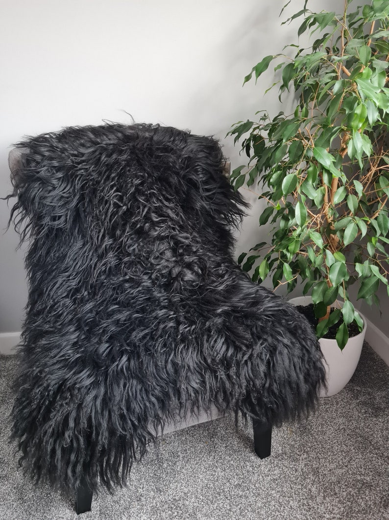 May include: A long-haired black sheepskin rug draped over a chair. The texture is thick, shaggy, and curly, covering the seat and backrest of the chair completely. The deep black fur has a natural, voluminous appearance with varied hair lengths.