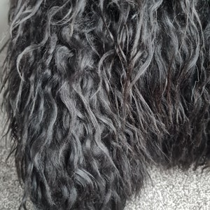 May include: A dense, black, long-haired textile material with a wavy, curly texture. The fibers are dark and layered, creating a voluminous appearance with deep shadows between the individual strands of the dark, textured, shaggy material.