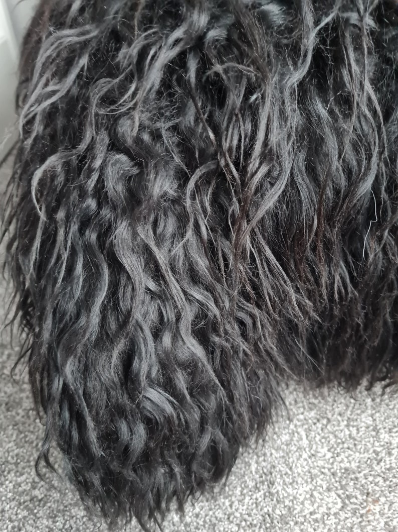May include: A dense, black, long-haired textile material with a wavy, curly texture. The fibers are dark and layered, creating a voluminous appearance with deep shadows between the individual strands of the dark, textured, shaggy material.